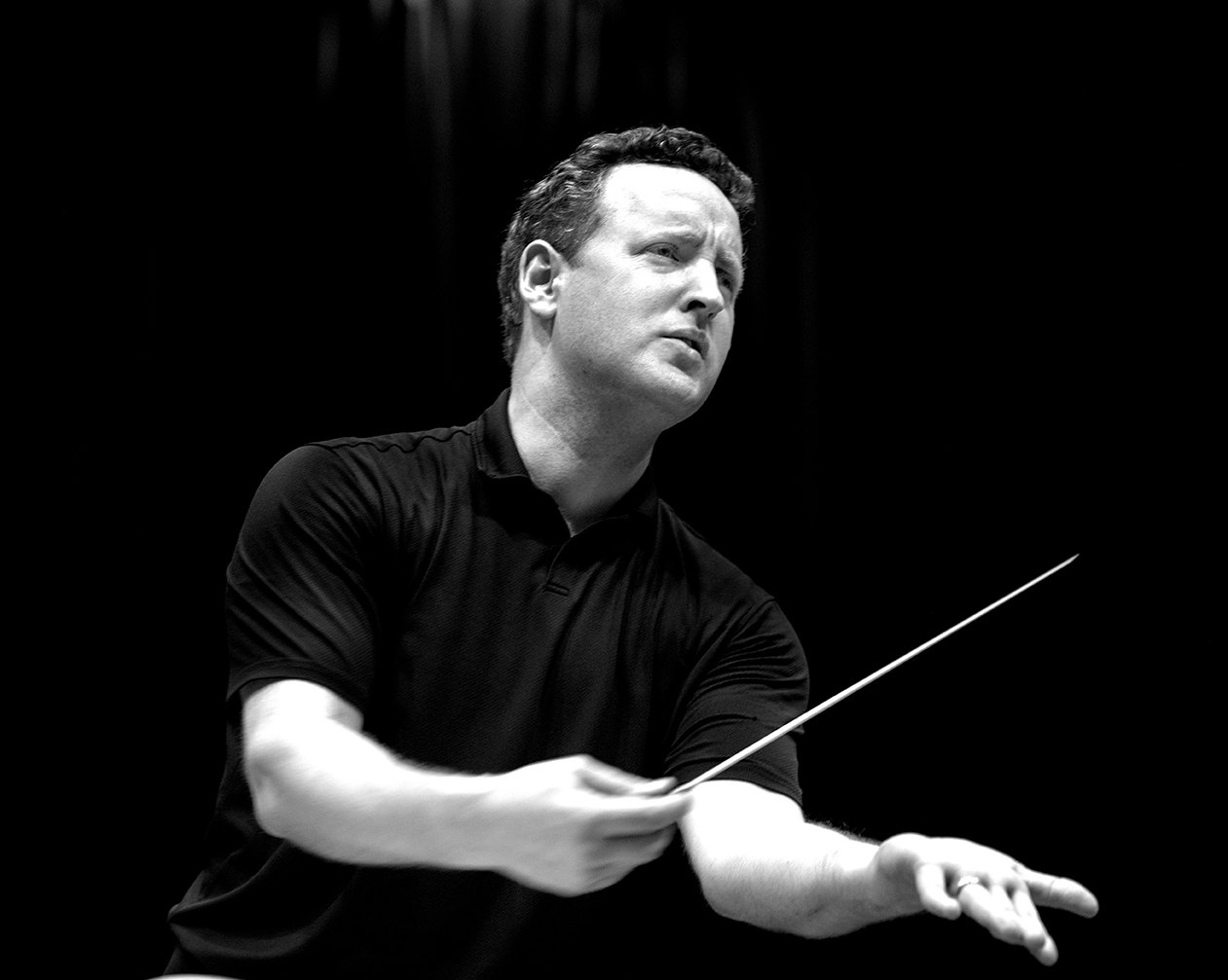 Michael Francis conducting in a black shirt, baton extended, gazing upward