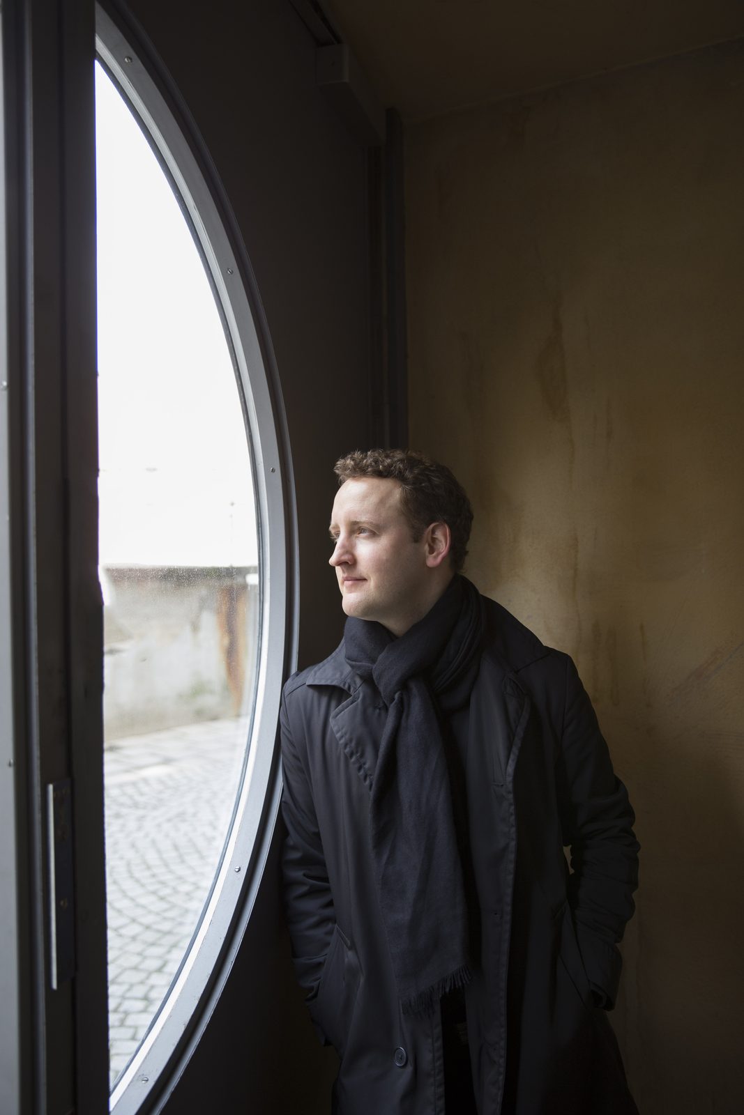Michael Francis by a tall round window in a coat and scarf